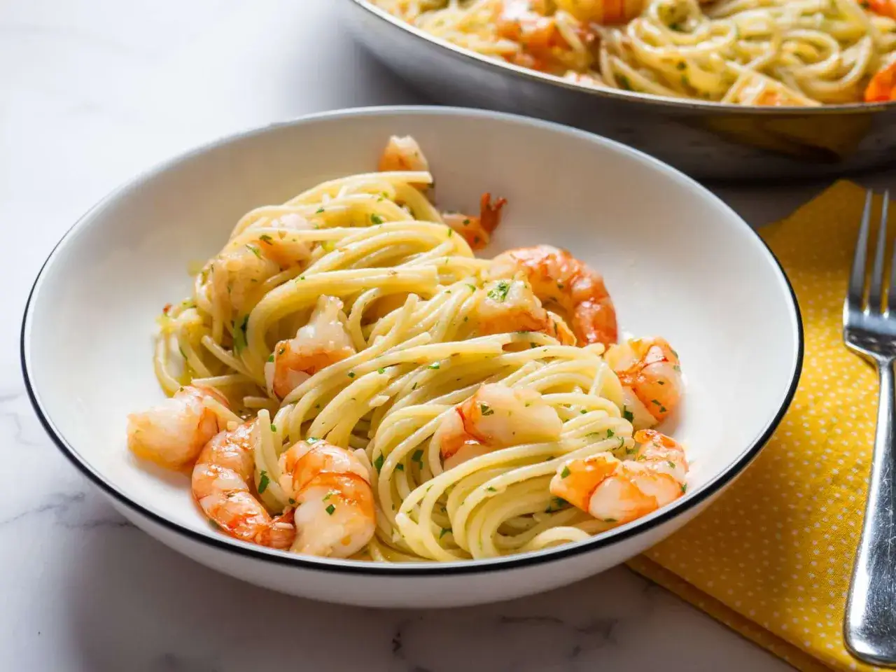 Ein Teller mit köstlichem Spaghetti mit Garnelen, garniert mit Petersilie. Im Hintergrund eine Pfanne mit demselben Gericht.