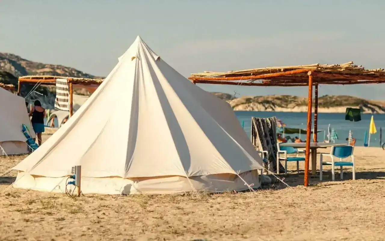 Piękne kempingi w Grecji nad morzem. Biały namiot na plaży, stół z krzesłami pod zadaszeniem, w tle woda i wyspy.