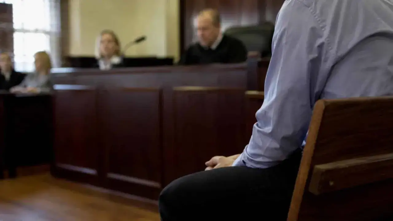 W sali sądowej mężczyzna w jasnej koszuli czeka na werdykt, mając nadzieję na wykupienie się od wyroku. Sędziowie w tle.