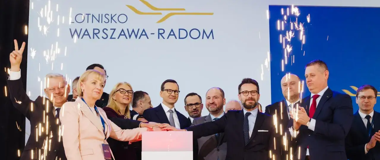 Uroczyste otwarcie lotniska Warszawa-Radom. Grupa osób z premierem na czele naciska przycisk, a wokół nich sypią się sztuczne ognie.
