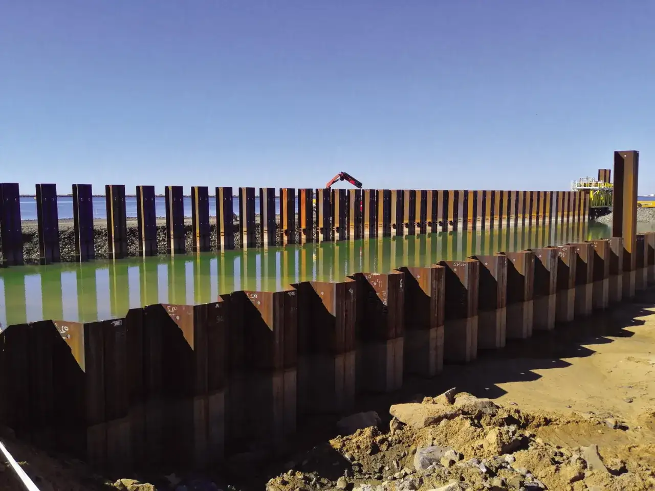 Una barrera de **perfiles laminados de acero** se alza sobre el agua verde, reflejando el cielo azul.