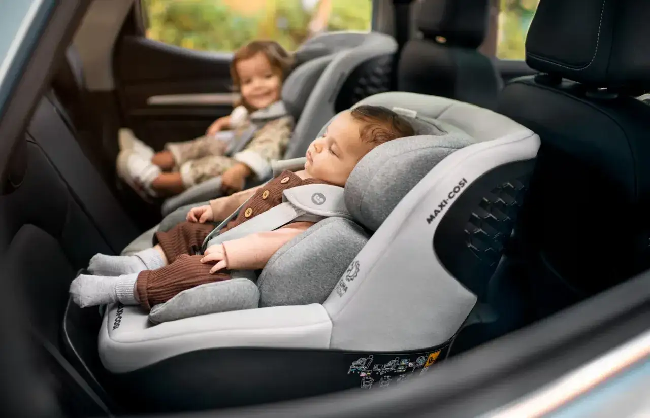 Bebé durmiendo en una silla de coche Maxi-Cosi, con otro niño sonriendo detrás. Seguridad y comodidad para los más pequeños en cada viaje.
