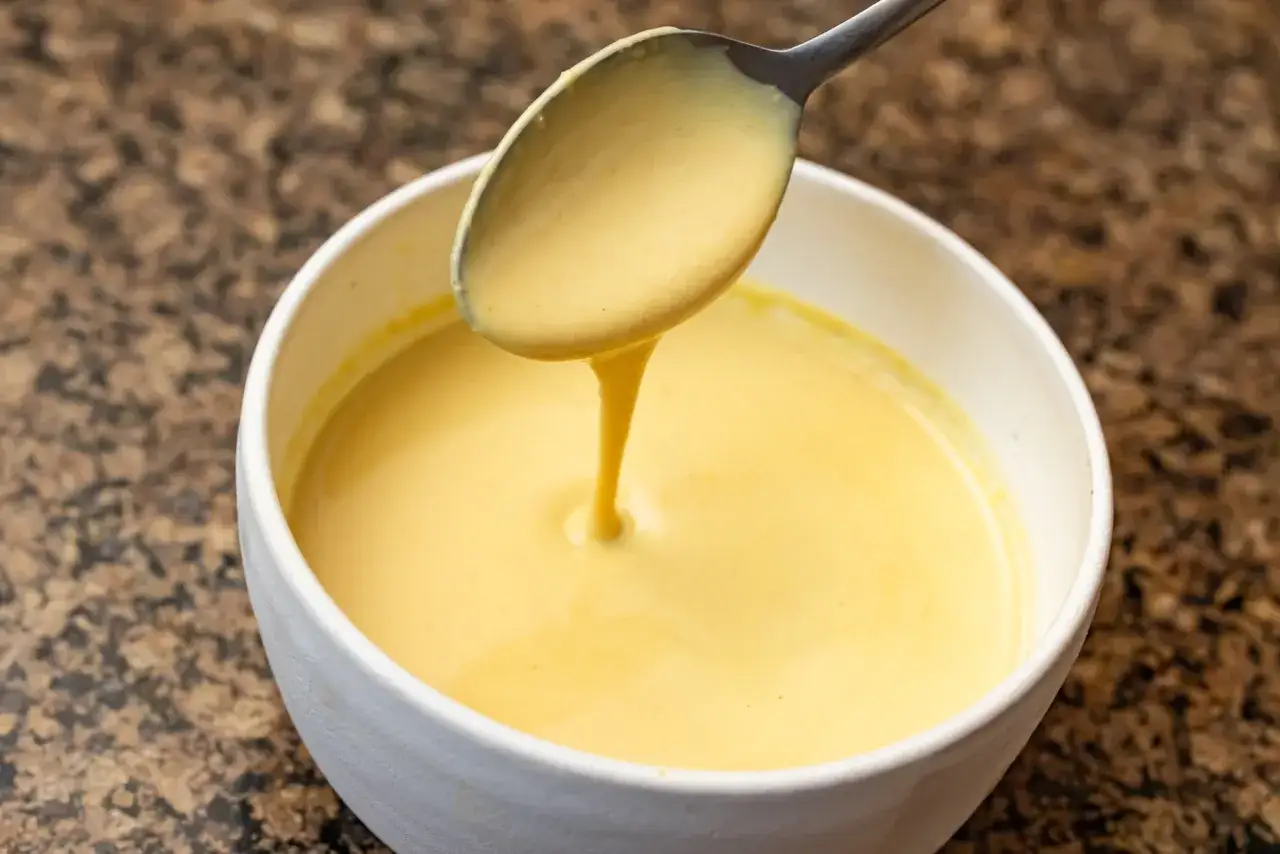 Cremige vegane hollandaise tropft von einem L&ouml;ffel in eine wei&szlig;e Sch&uuml;ssel.
