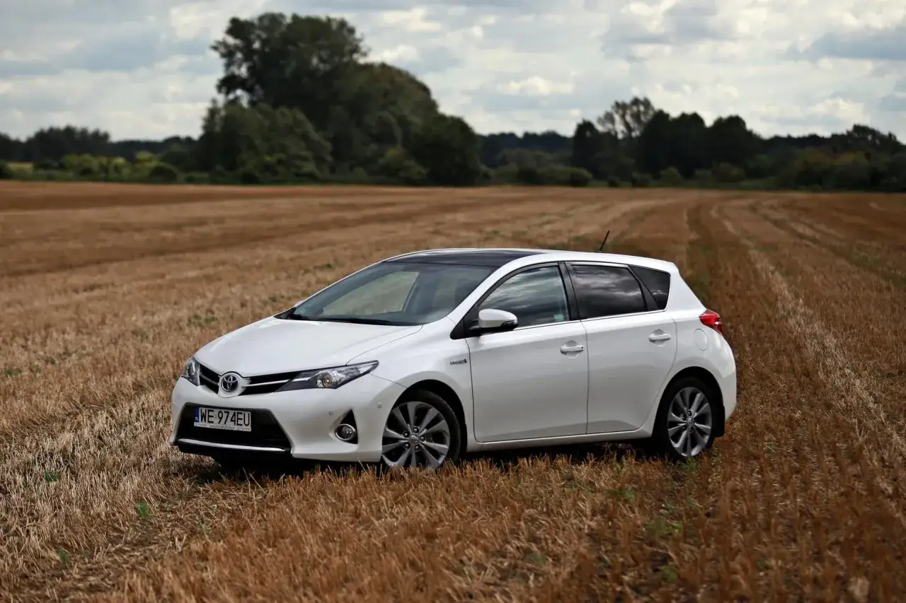 Biała Toyota Auris Hybrid na polu po żniwach. Ten model, jak każdy samochód hybrydowy, posiada specjalny akumulator, który zasila silnik elektryczny.