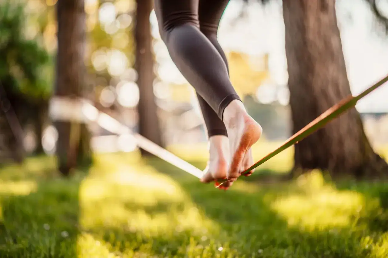 Chodzenie po taśmie to slackline. Stopy balansują na taśmie rozwieszonej między drzewami, w słonecznym lesie.