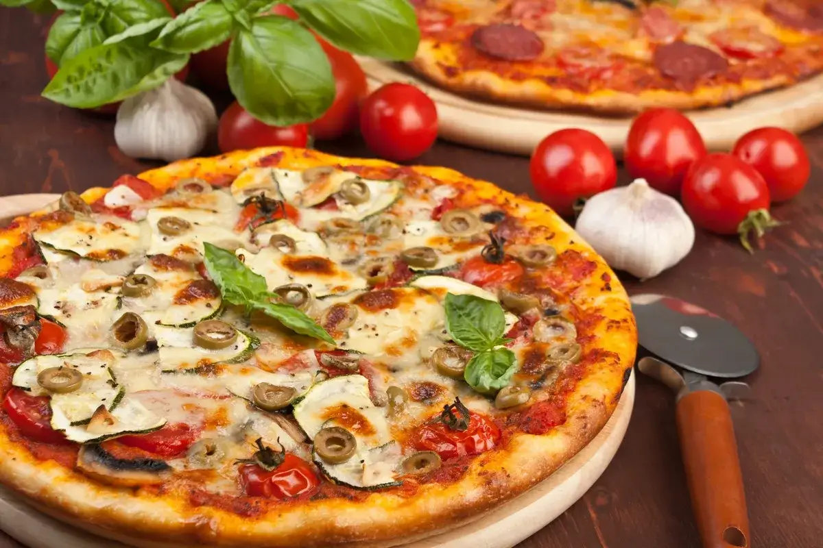 Pyszna pizza z cukinią i oliwkami, idealna do przygotowania pieczarek do pizzy. Obok świeże pomidory i czosnek.