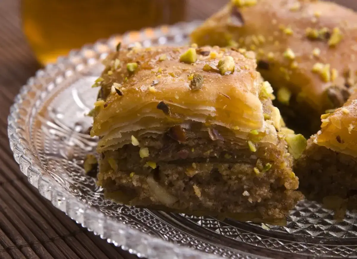 Słodka baklava, co to jest? Warstwy ciasta filo z orzechami i syropem, posypana pistacjami, na szklanym talerzyku.