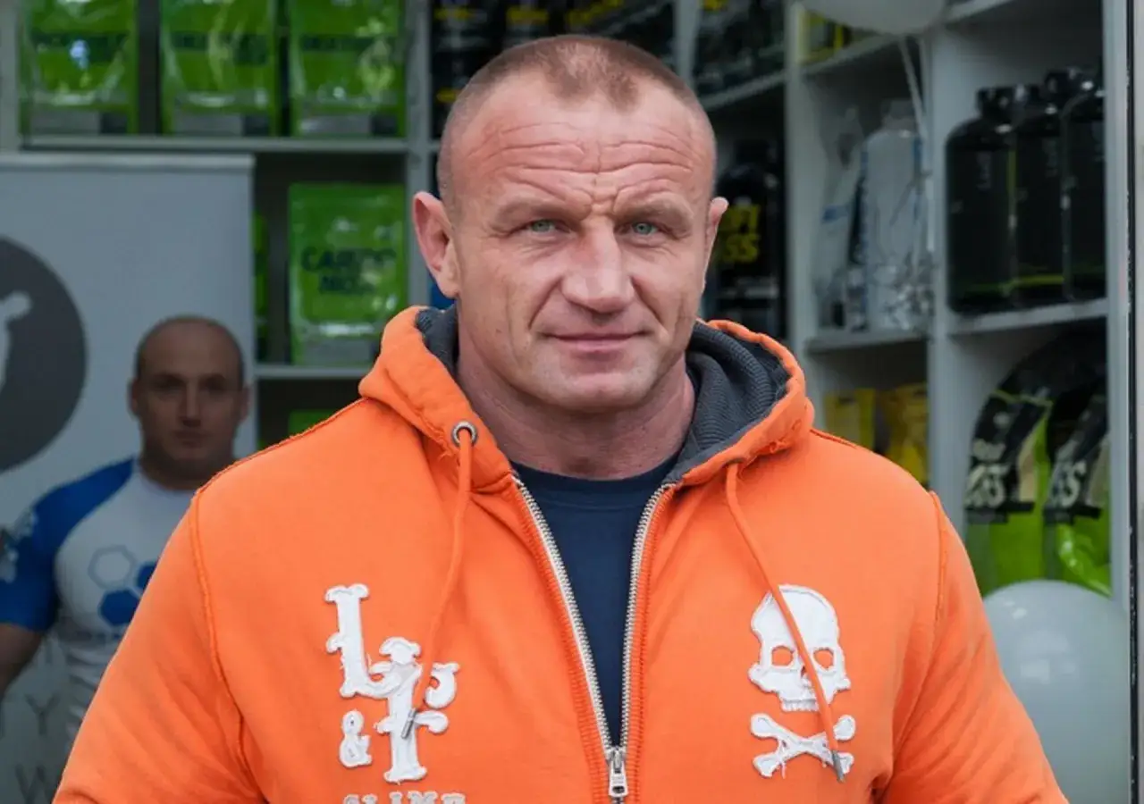 Mocny mężczyzna w pomarańczowej bluzie z kapturem, z czaszką i skrzyżowanymi piszczelami. Ile lat ma Pudzian? Wiek to tylko liczba dla tego sportowca.