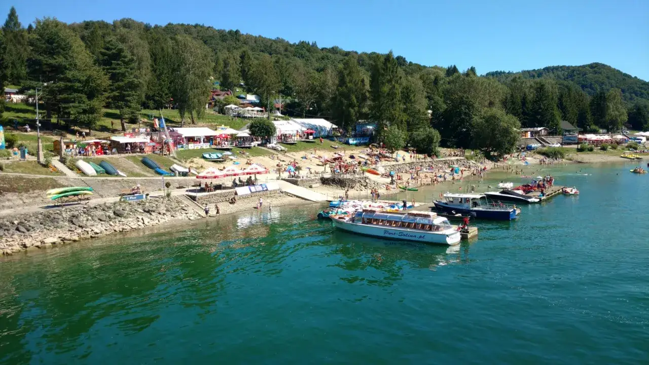 Tłoczna solina plaża z ludźmi kąpiącymi się i opalającymi. W wodzie pływają łodzie i rowerki wodne.
