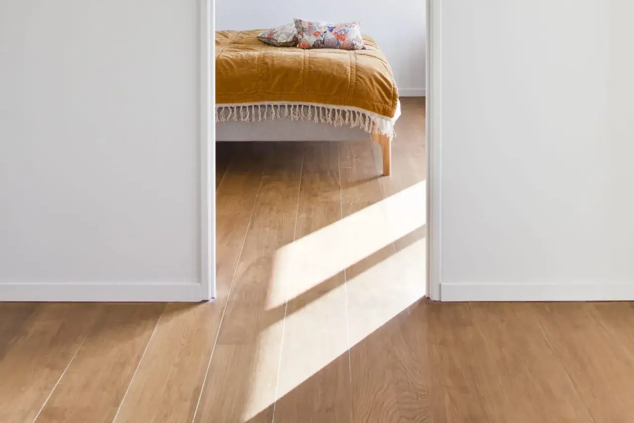 Vue d'une chambre avec un lit couvert d'une couverture ocre. Le sens pose parquet chambre est mis en valeur par des rayons de soleil traversant l'ouverture.