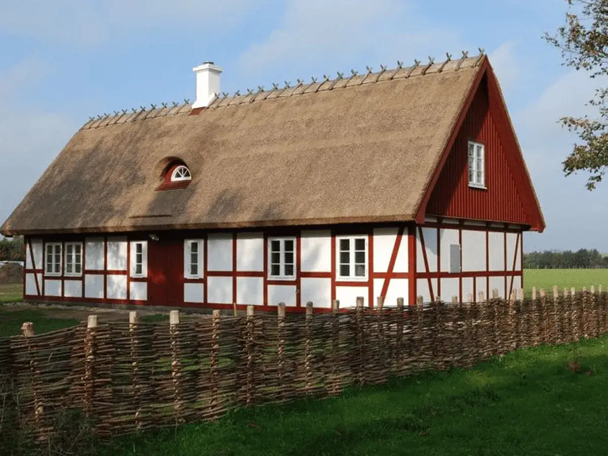 Urokliwy dom szachulcowy z czerwonymi elementami i strzechą na dachu, otoczony plecionym płotem.