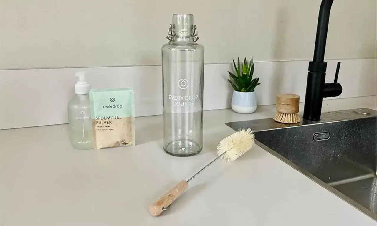 Alles bereit f&uuml;r die Edelstahl Trinkflasche reinigen: Sp&uuml;lmittel, Pulver und eine Flaschenb&uuml;rste liegen neben der Glasflasche.