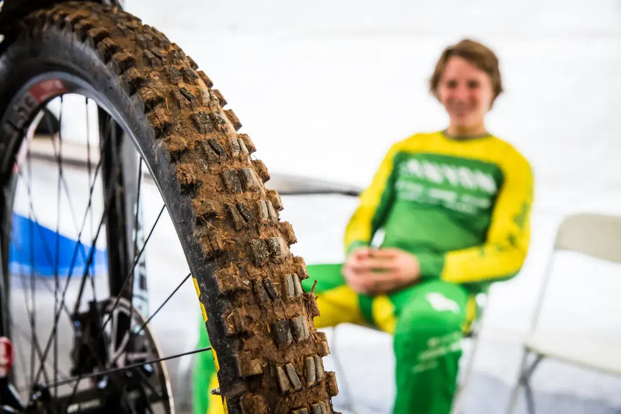 Close-up of a muddy Maxxis Minion DHF tire, ready for a downhill review. A smiling rider in a green and yellow jersey is blurred in the background.