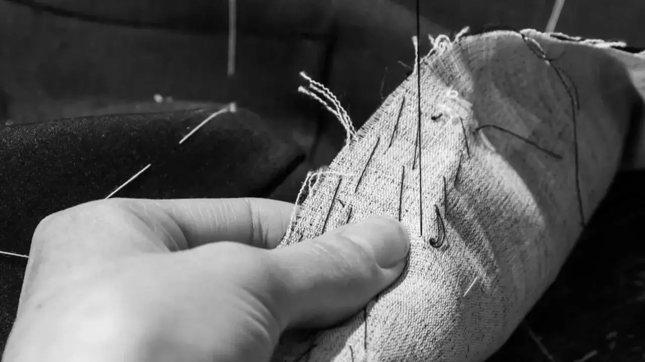 A hand pins fabric for suit construction. Needles and threads are visible, highlighting the meticulous tailoring process.