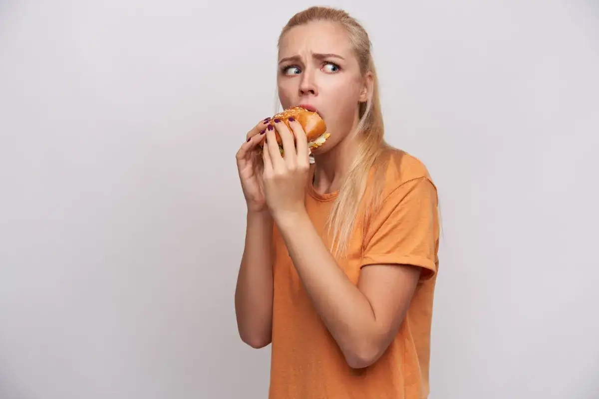 Blondynka w pomarańczowej koszulce z przerażeniem w oczach je hamburgera, walcząc z kompulsywnym objadaniem się.