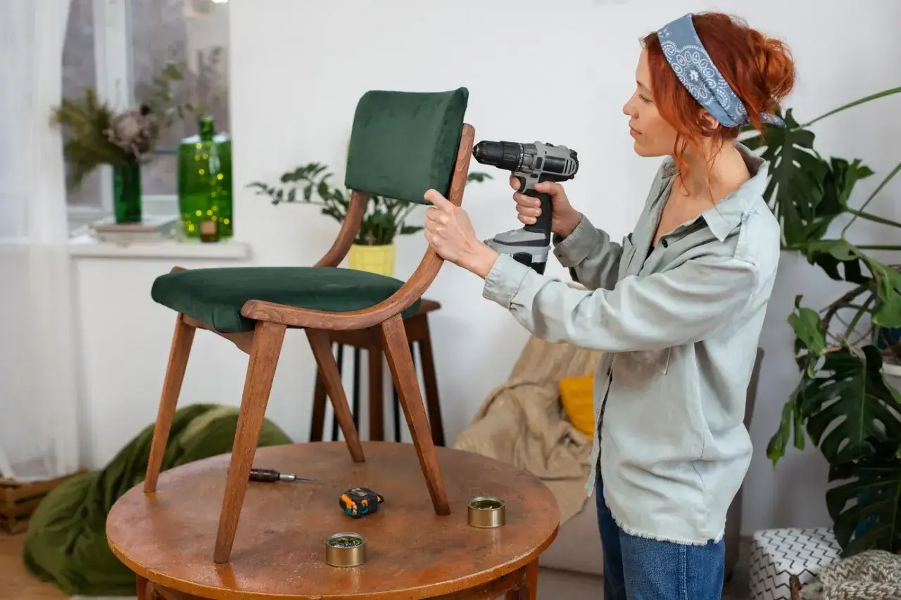 Woman uses a drill to reupholster a vintage chair with green velvet. Tools and supplies are on the table.