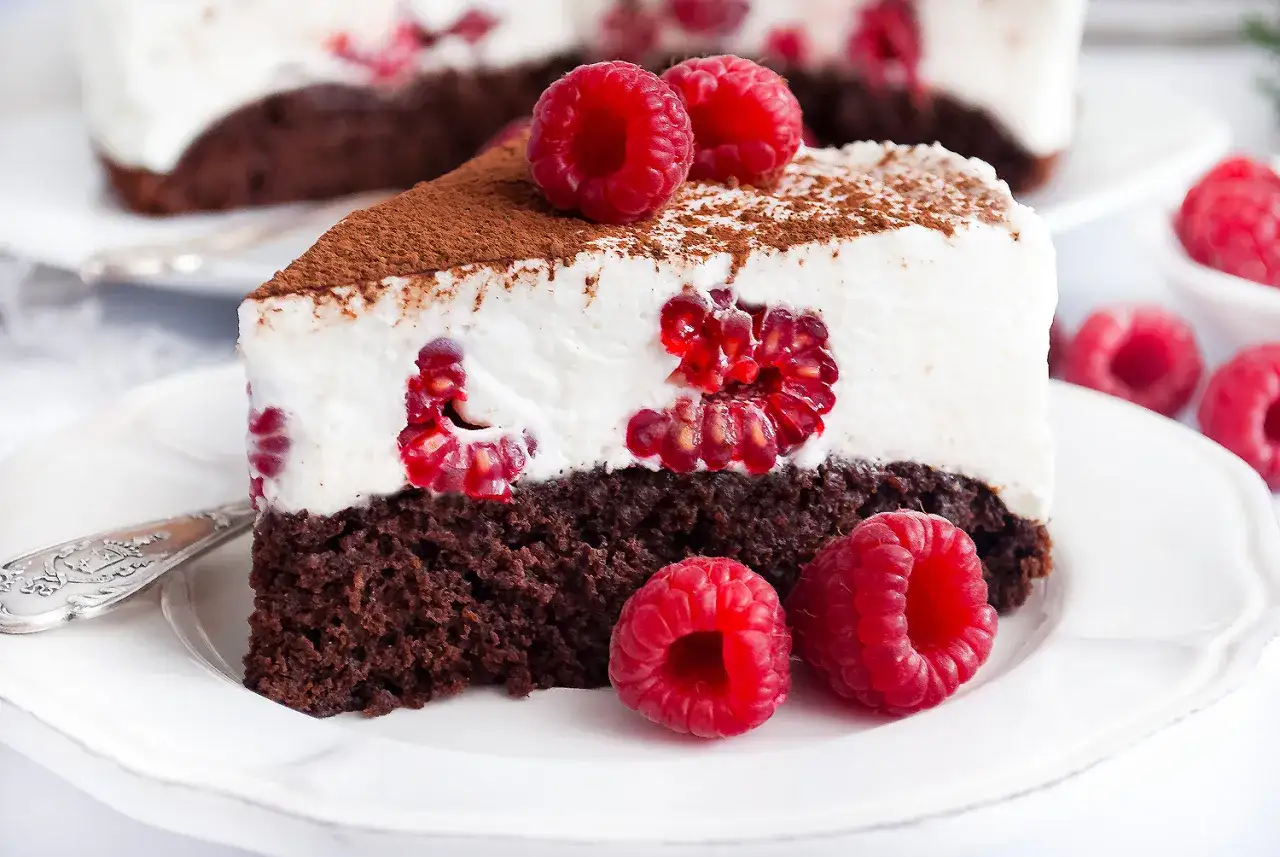 Pyszny brownie z malinami, udekorowany bitą śmietaną i kakao, kusi świeżymi malinami.