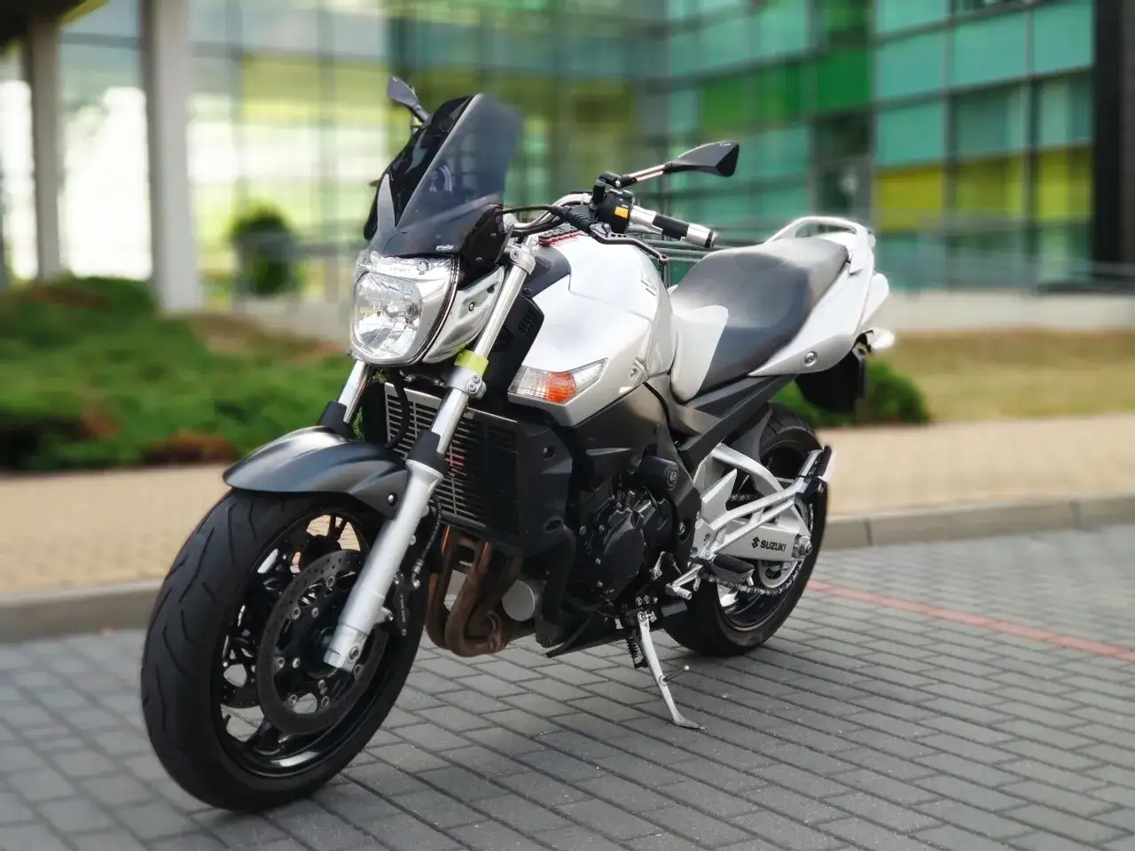 Srebrny motocykl Suzuki GSR600 z czarną szybką i siedzeniem, zaparkowany na chodniku przed nowoczesnym budynkiem.