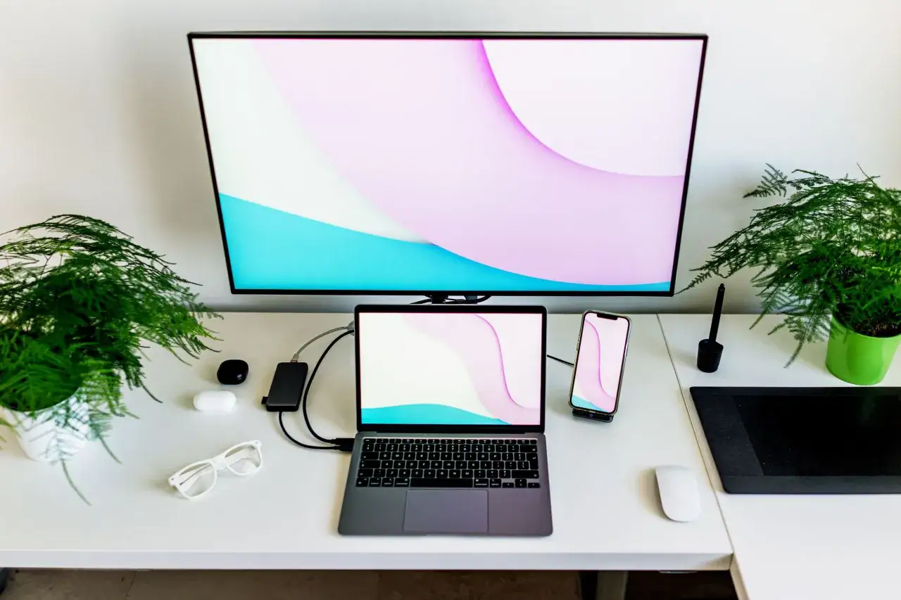 Ein moderner Arbeitsplatz mit einem Laptop, der mit einem gro&szlig;en Bildschirm verbunden ist. Pflanzen und ein Smartphone erg&auml;nzen das Bild.