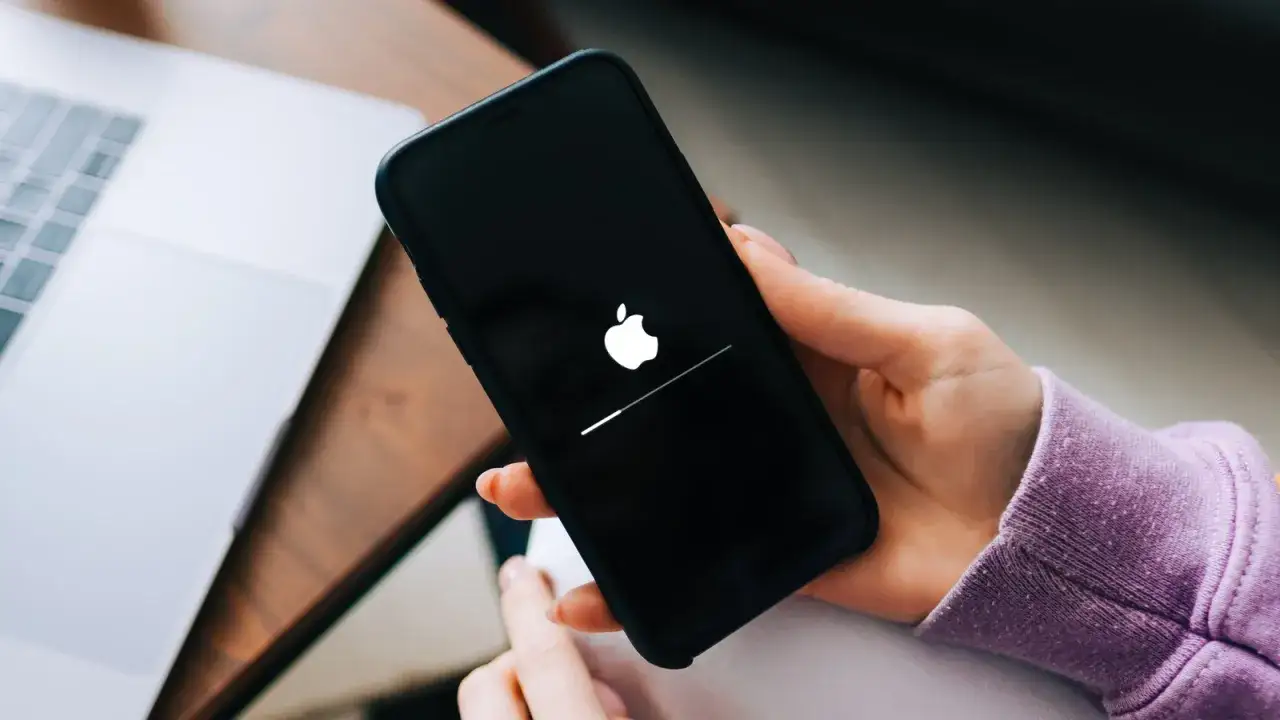 Dłoń trzyma telefon, na którym widnieje logo Apple i pasek postępu. Wygląda na to, że iPhone nie chce się włączyć.