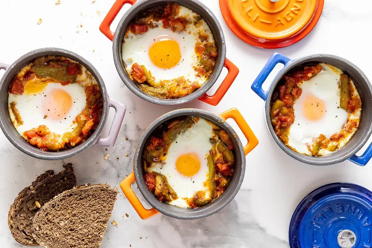 Quattro cocotte colorate con uova in camicia su un letto di pomodoro e peperoni. Un'idea per ricette con uova gustose e veloci.