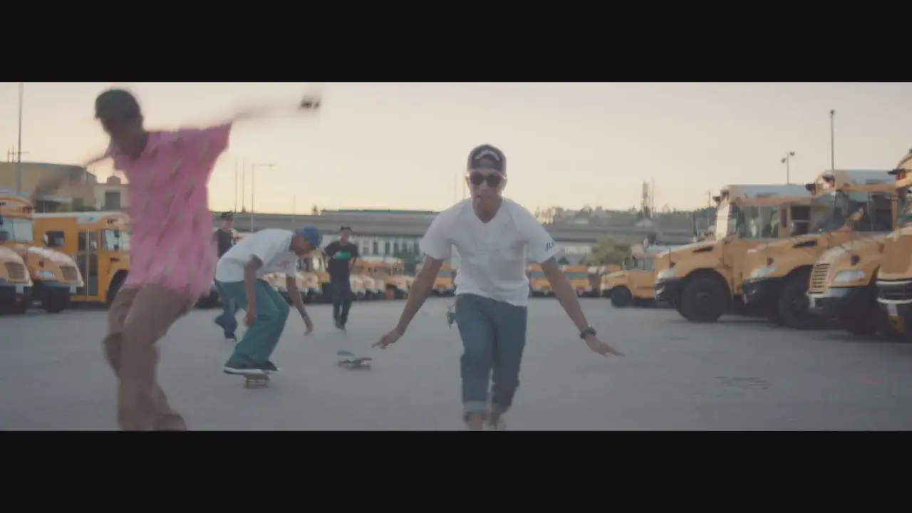 Młodzi ludzie jeżdżą na deskorolkach przed rzędem żółtych autobusów. Pharrell Williams, szczęśliwy, w różowej koszulce, w ruchu.