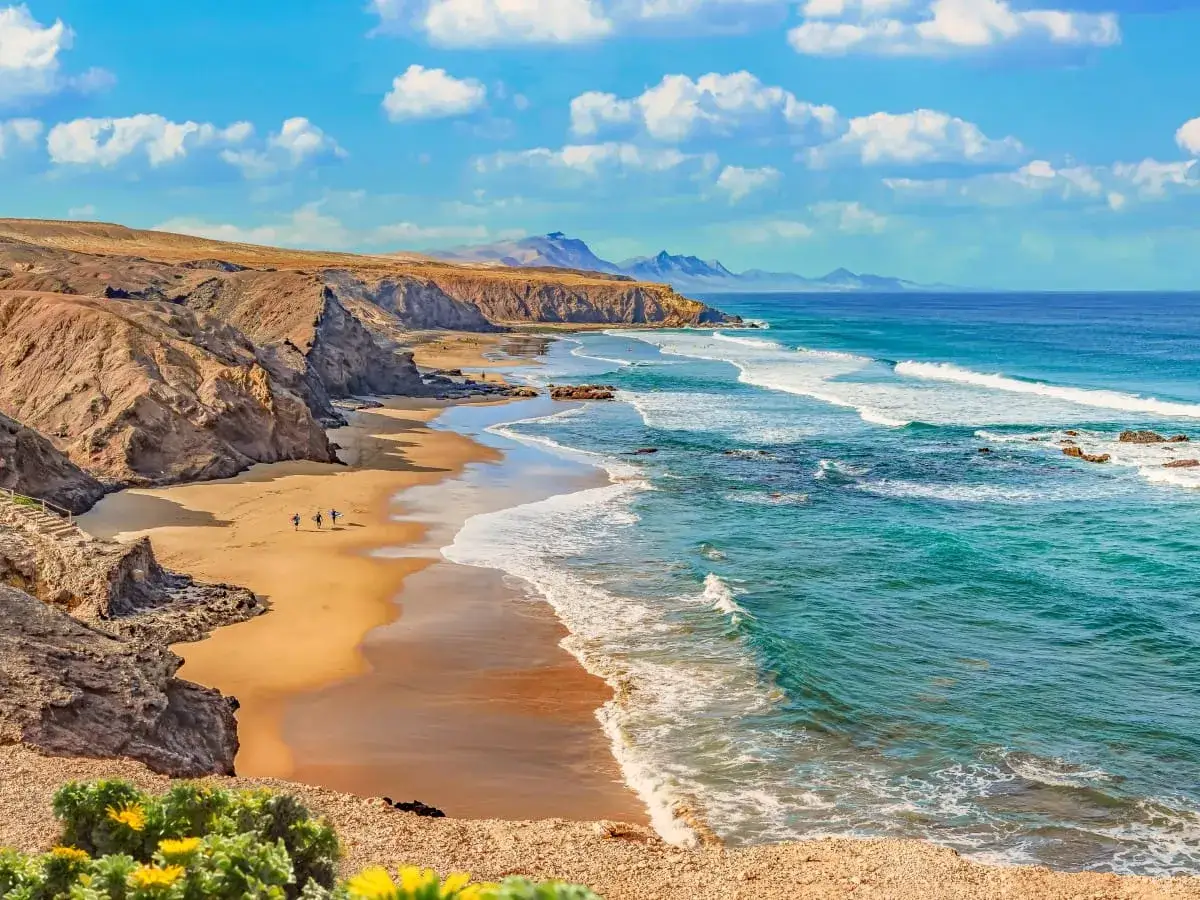 Sonnenbeschienener Surfer Strand Fuerteventura mit türkisfarbenem Meer, Wellen und felsigen Klippen. Drei Surfer laufen am Sandstrand entlang.