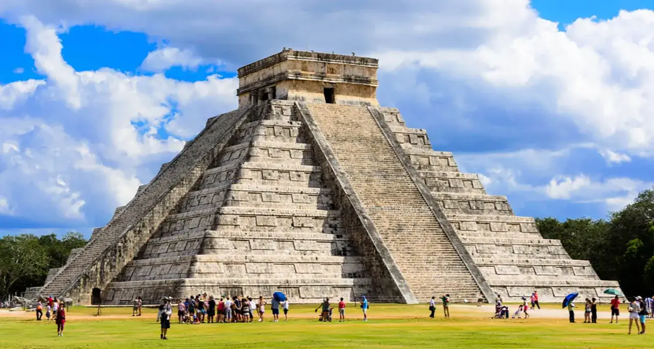 La piramide di Kukulkan a Chichen Itza, con turisti che ammirano la sua maestosità sotto un cielo azzurro e nuvoloso.