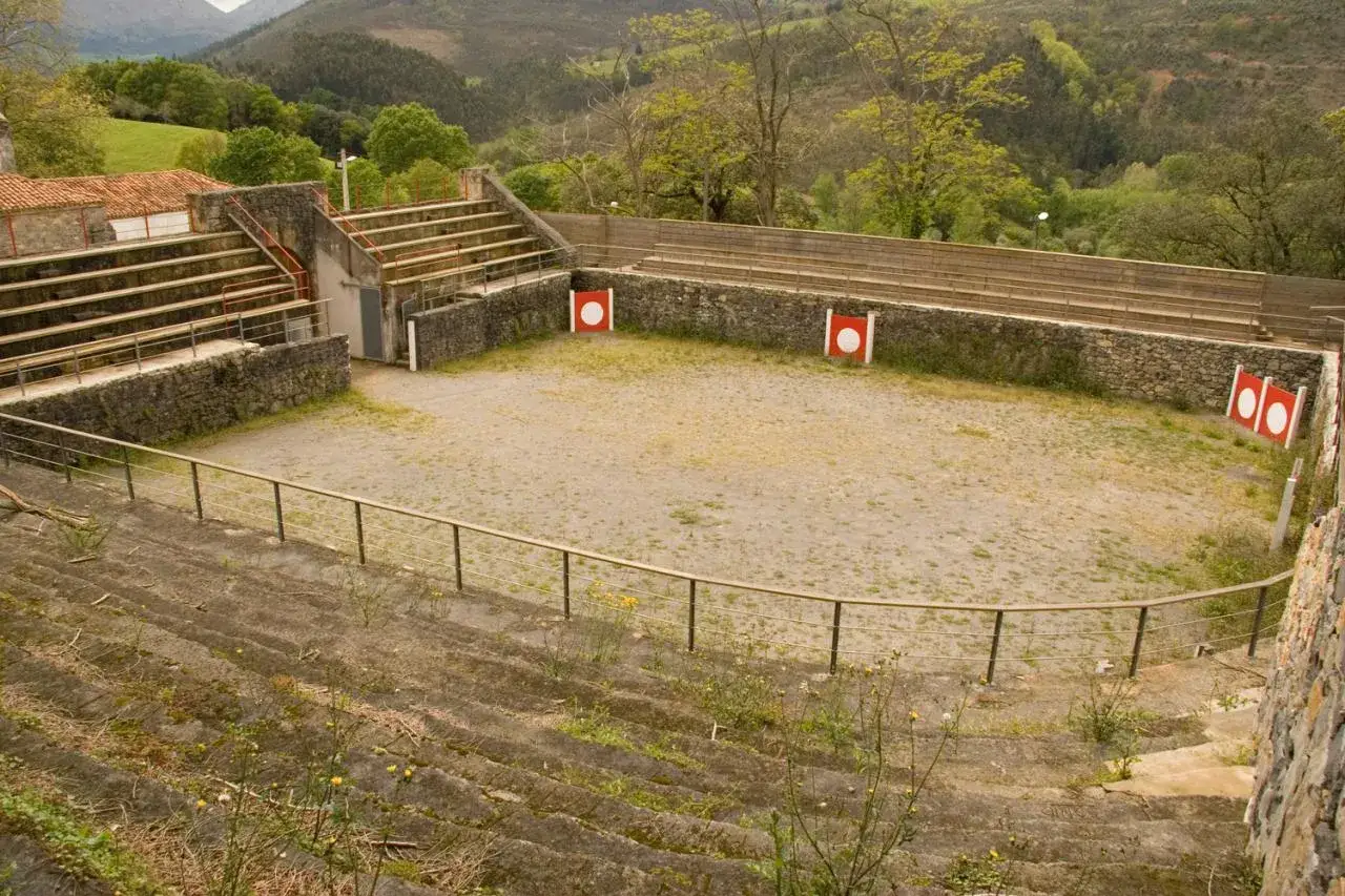 Plaza de toros cuadrada en Espa&ntilde;a, con gradas de piedra y arena en el ruedo. Rodeada de vegetaci&oacute;n y colinas.