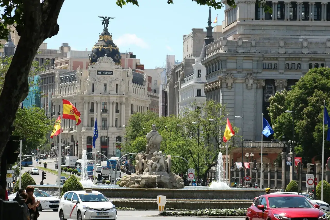 Edificio Metropolis en Madrid, con banderas españolas ondeando. El tráfico fluye, sugiriendo la actividad de negocios millonarios en esta vibrante ciudad.