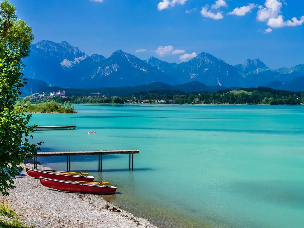 T&uuml;rkisfarbener See mit zwei roten Ruderbooten am Ufer, bereit f&uuml;r eine Fahrt. Im Hintergrund Berge und eine kleine Stadt. Perfekt zum Schwimmen.