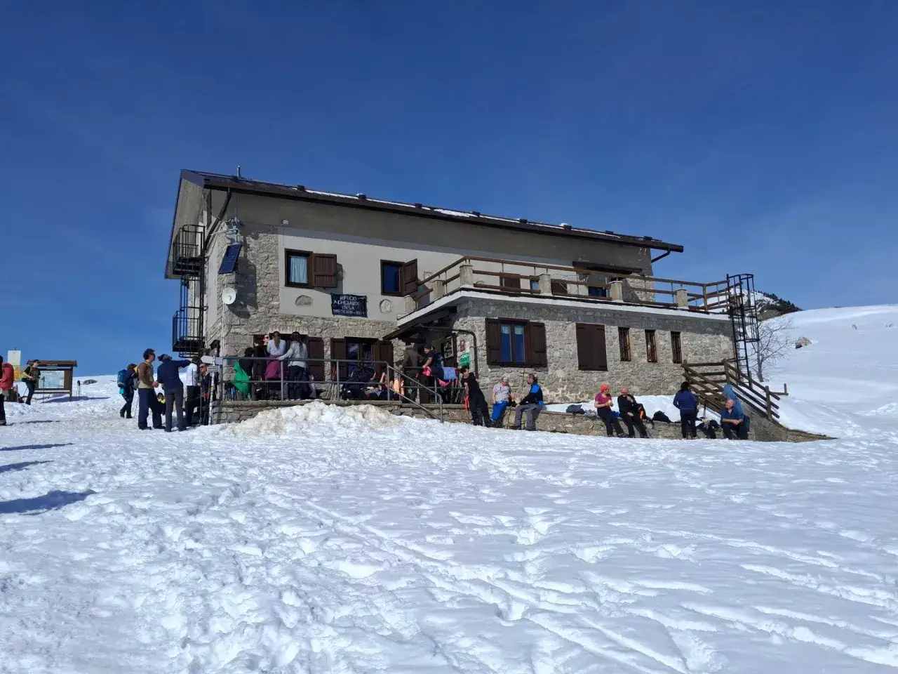 Persone riposano sulla neve davanti al rifugio Gherardi, un edificio in pietra con tetto spiovente sotto un cielo azzurro.