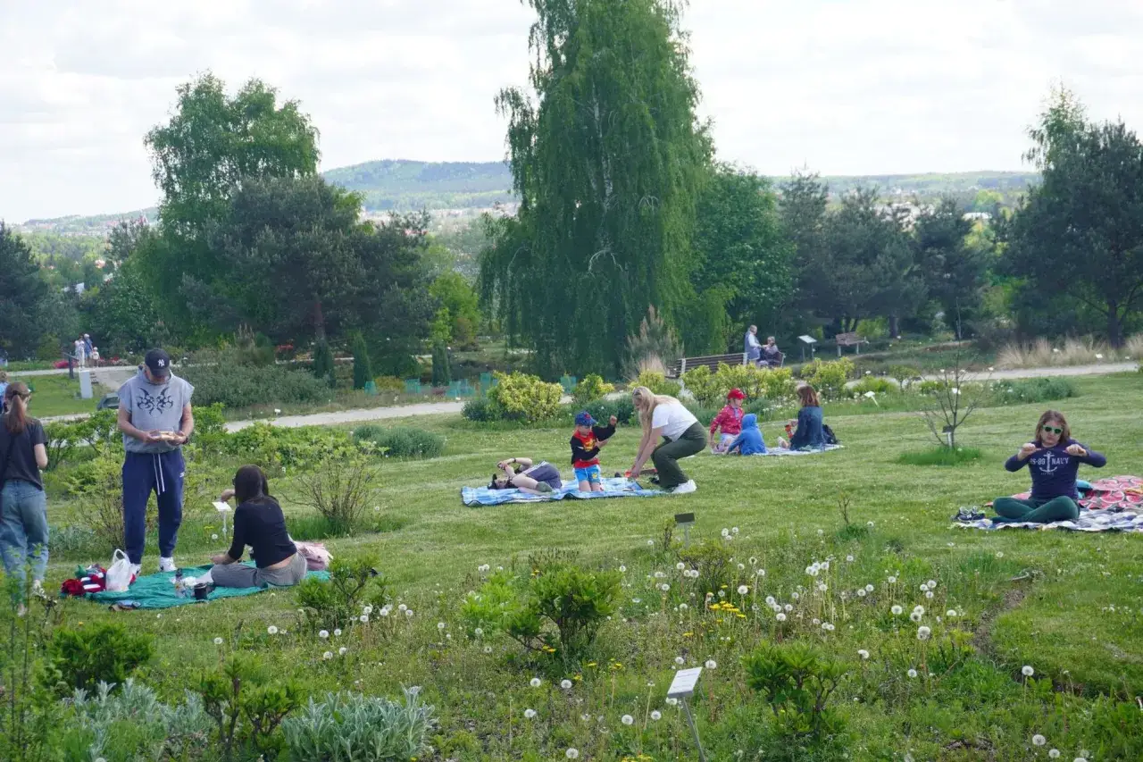 Ludzie piknikują w ogrodzie botanicznym, jednym z romantycznych miejsc w Kielcach. Dzieci bawią się na trawie, dorośli odpoczywają.