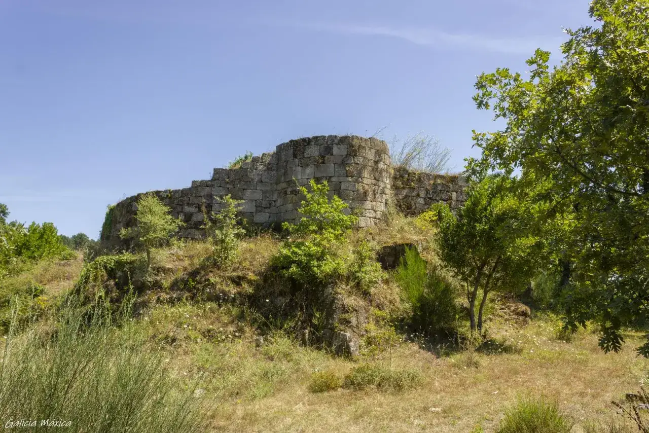 Ruinas de una antigua **peroxa** de piedra, rodeada de vegetación y bajo un cielo azul.