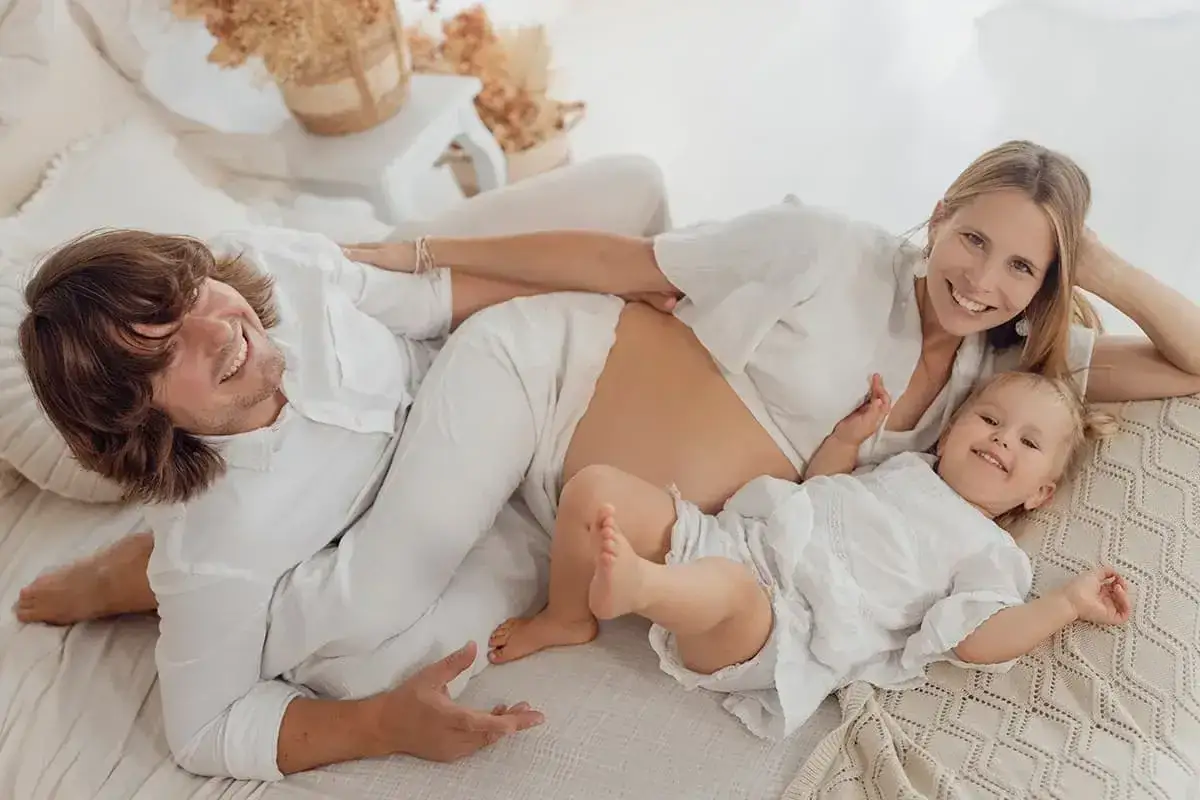 Szczęśliwa rodzina podczas sesji ciążowej z mężem. Uśmiechnięta mama z brzuszkiem, tata i mała córeczka leżą razem na łóżku.