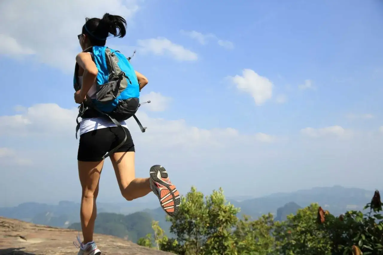 Donna con zaino blu corre in montagna, godendosi il panorama mozzafiato sotto un cielo azzurro con nuvole bianche.