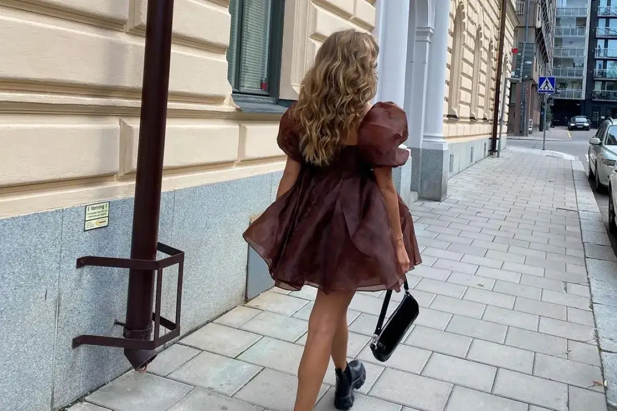 Kobieta w brązowej sukience z bufiastymi rękawami i czarną torebką idzie chodnikiem. TPO.
