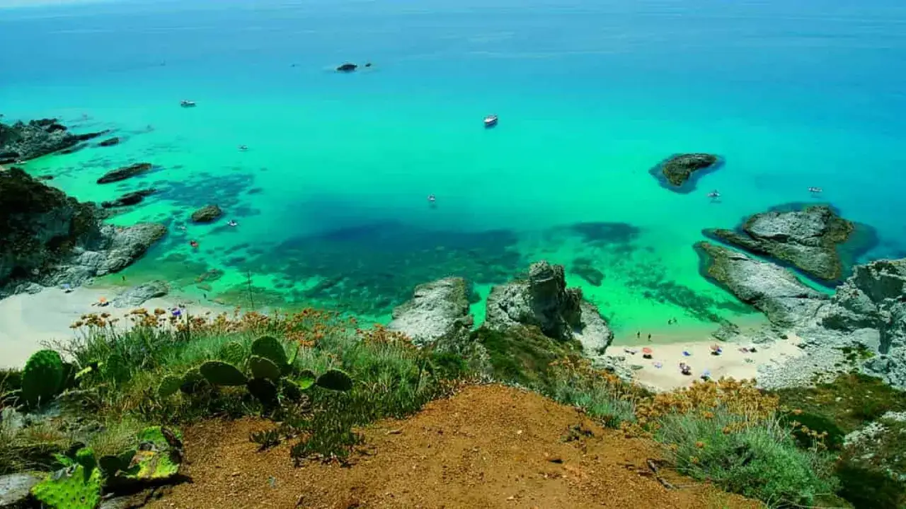 Vista dall'alto di una baia con acque cristalline e sabbia dorata, tipica delle **calabria spiagge**. Rocce affiorano dal mare turchese, mentre piccole imbarcazioni solcano la superficie.