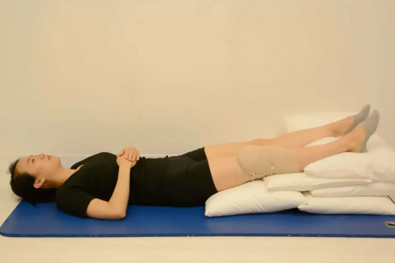 Donna sdraiata su un tappetino blu, gambe sollevate su cuscini, con una fascia compressiva sul ginocchio per alleviare i **caviglie gonfie**.