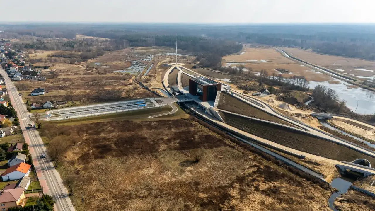Nowoczesny budynek muzeum Bitwy Warszawskiej z charakterystycznym obeliskiem na tle krajobrazu z drogą, domami i terenami podmokłymi.