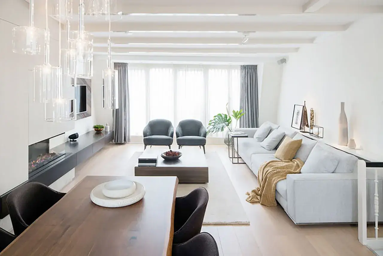Elegante sal&oacute;n comedor dos ambientes con chimenea, sof&aacute;s grises, mesa de centro y l&aacute;mparas colgantes de cristal.