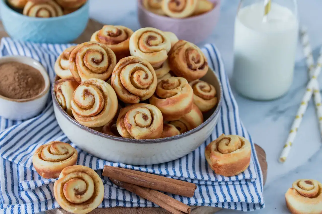 Sch&uuml;ssel voller goldbrauner mini zimtschnecken, daneben Zimtstangen und ein Glas Milch.