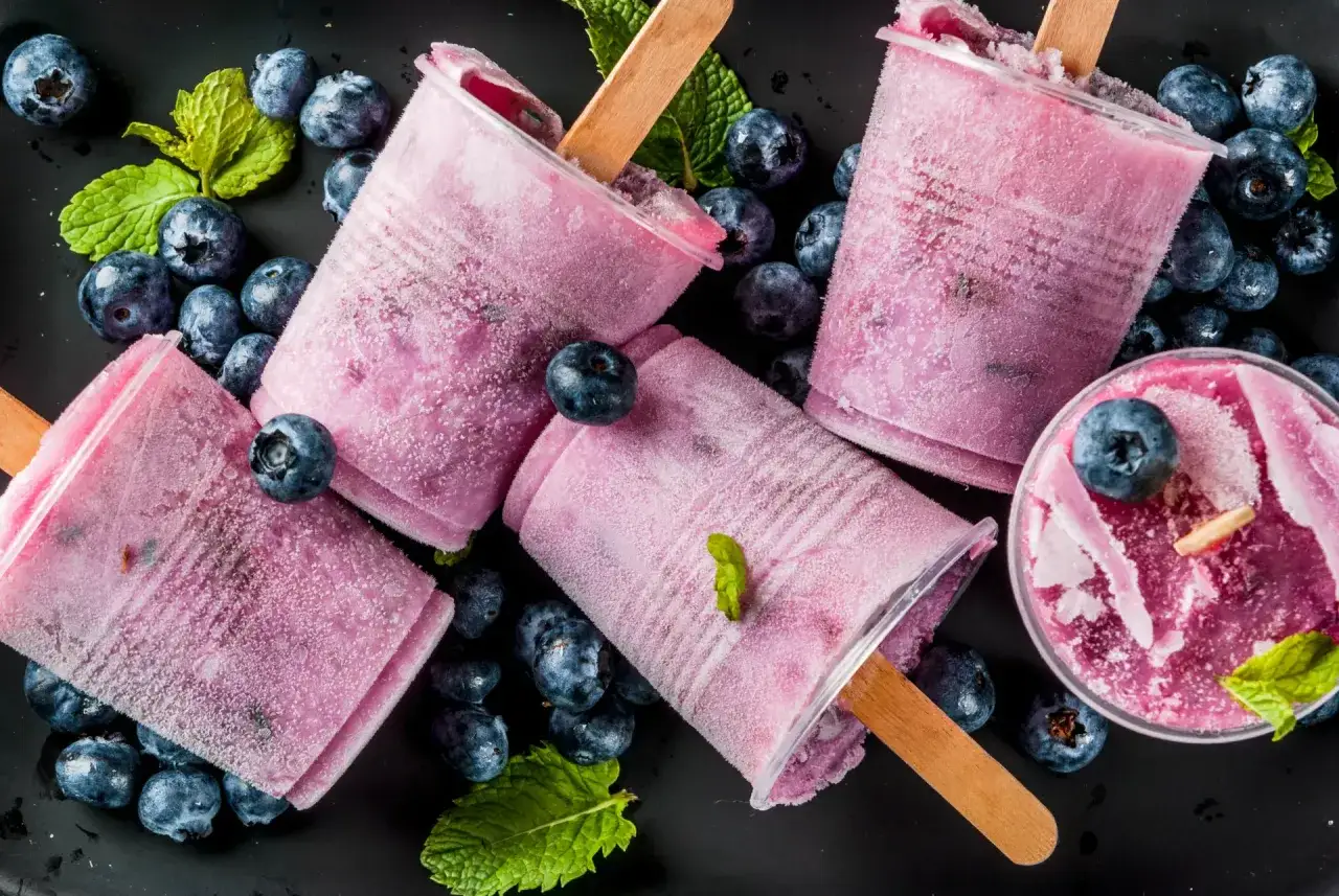 Przepisy na zdrowe lody jagodowe. Lody na patyku i w kubeczku, otoczone świeżymi jagodami i listkami mięty.