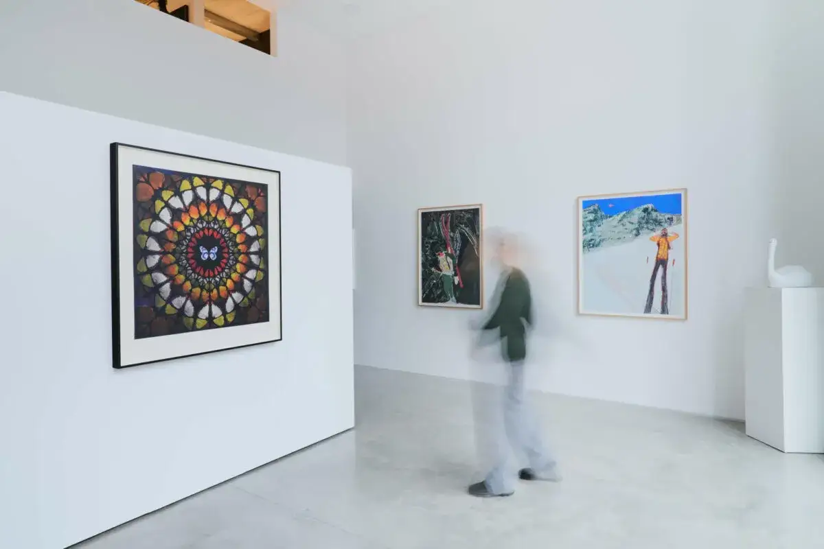 A gallery visitor admires a vibrant red chip art mandala with a butterfly at its center, alongside other paintings.