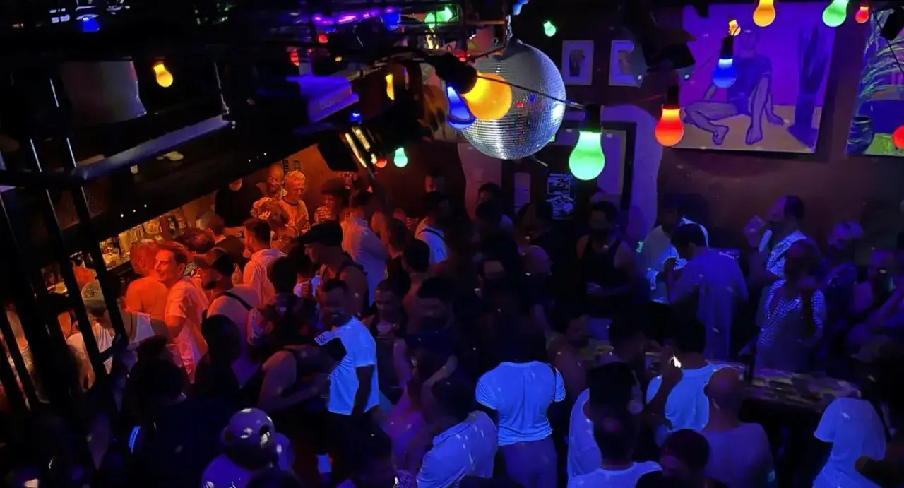 Ambiance festive dans un bar gay, avec une foule animée sous des lumières colorées et une boule disco.