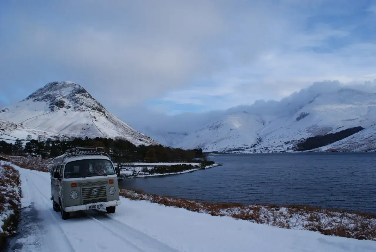 Ein Camper f&auml;hrt auf einer verschneiten Stra&szlig;e an einem See entlang. Im Hintergrund erheben sich schneebedeckte Berge. Eine malerische England Landschaft im Winter.