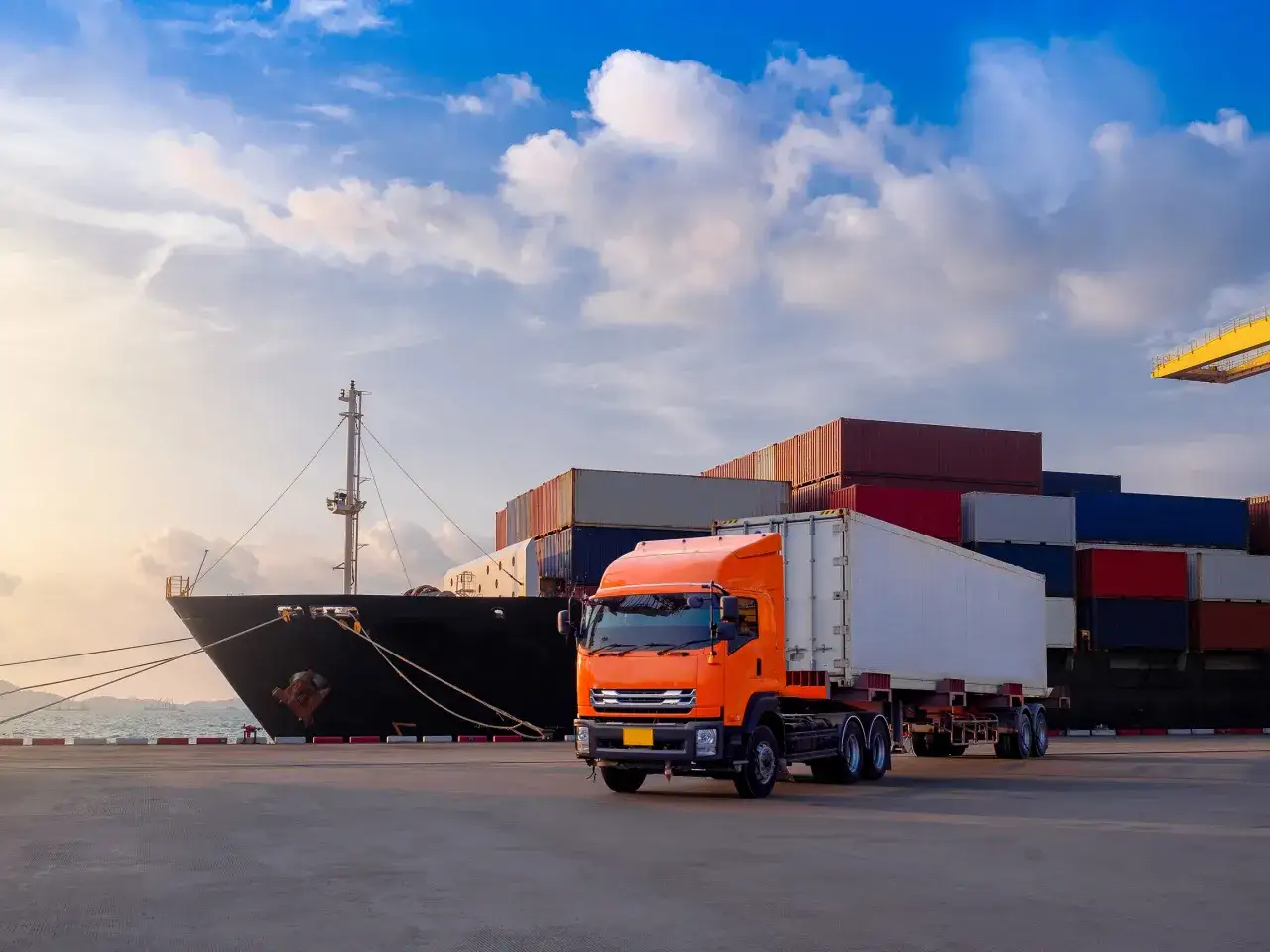 Orange Sattelzug wartet auf Fracht neben Containerschiff im Hafen. Der LKW ist Teil des merchant haulage Prozesses.