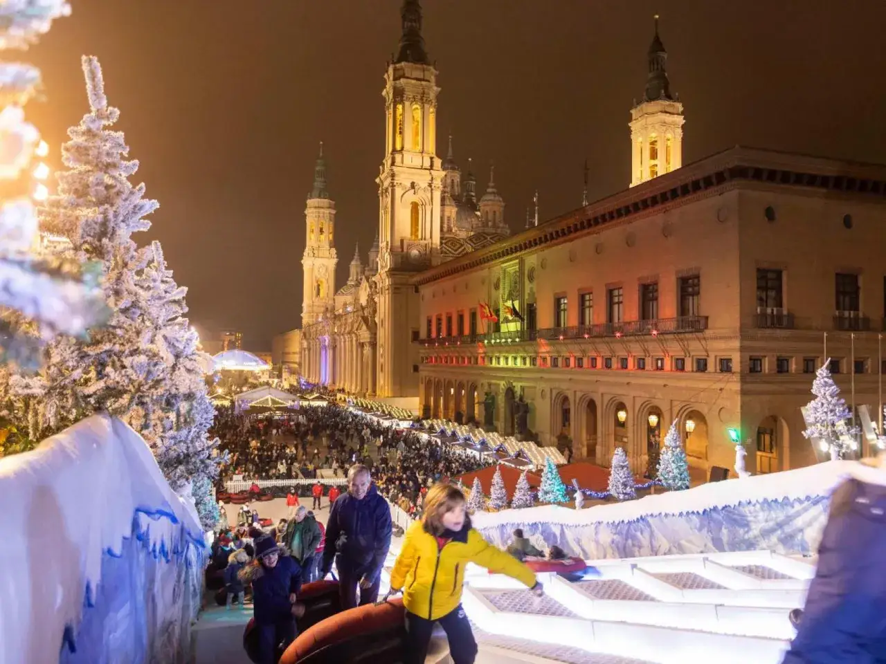 Niños disfrutan de un tobogán nevado en el mercadillo navideño de Zaragoza, con la Basílica del Pilar iluminada de fondo.