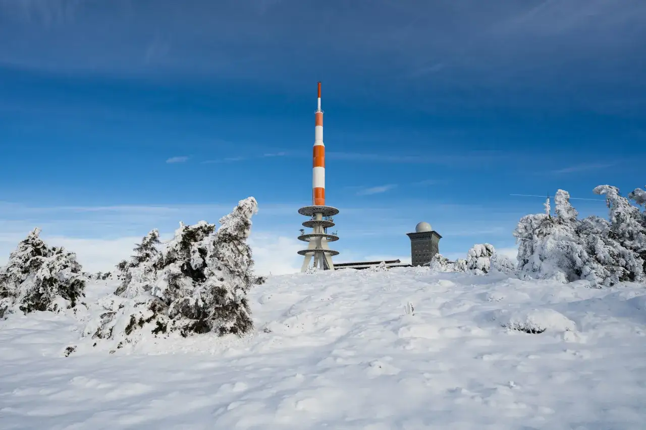 Winter am Brocken: Ein rot-weißer Sendeturm und ein Gebäude ragen aus einer verschneiten Landschaft mit eisverkrusteten Bäumen unter blauem Himmel.