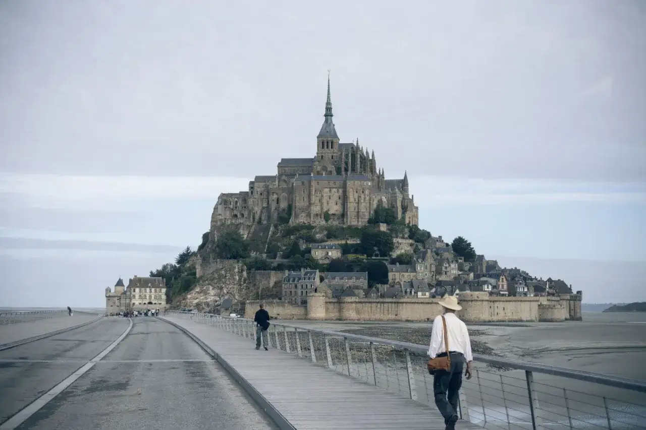 En man med hatt g&aring;r p&aring; en g&aring;ngbro mot Mont Saint-Michel, en av Frankrikes mest ikoniska sev&auml;rdheter.